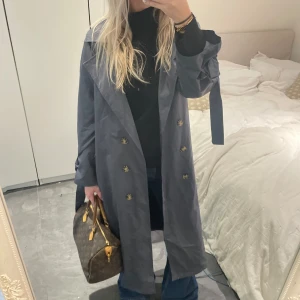 Grå trenchcoat med knappar - Snygg grå trenchcoat med knappar och bälte i midjan. Perfekt för höst och vår. Jackan har en klassisk design med långa ärmar och en stilren look.❣️❣️