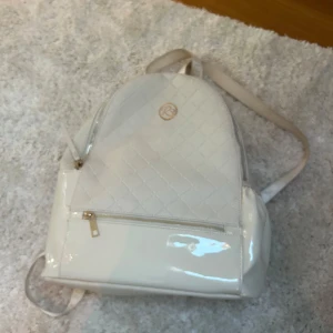 Vit ryggsäck från River Island - Snygg vit ryggsäck från River Island med quiltad design och dragkedja. Perfekt för att bära dina dagliga nödvändigheter med stil. Axelremmarna är justerbara och har märkets logga för en extra touch.