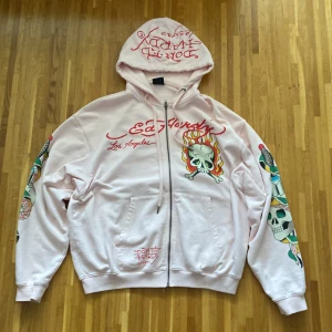 Rosa hoodie från Ed Hardy - Säljer en cool rosa hoodie från Ed Hardy med dragkedja och huva. Den har ett stort dödskalletryck med eldflammor på ryggen och ärmarna, samt Ed Hardy-logga på bröstet. Perfekt för dig som gillar unika och färgstarka plagg!