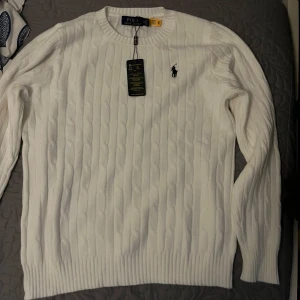 Vit kabelstickad tröja från Ralph Lauren - Säljer en stilren vit kabelstickad tröja från Ralph Lauren med deras ikoniska logga broderad på bröstet. Tröjan har en klassisk rund hals och långa ärmar, perfekt för en elegant och avslappnad look. Passar bra till både jeans och chinos.