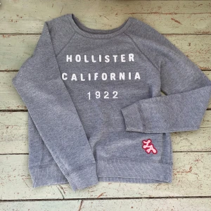 Grå sweatshirt från Hollister - Säljer en grå sweatshirt från Hollister med texten 'Hollister California 1922' tryckt på framsidan. Tröjan har långa ärmar och ribbade muddar vid ärmslut och nederkant. Perfekt för en avslappnad stil. 🩶