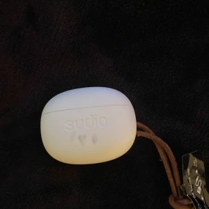 Trådlösa hörlurar från Sudio - Snygga trådlösa hörlurar från Sudio i ett stilrent vitt fodral. Hörlurarna har en elegant design med roséguldfärgade detaljer och kommer med ett praktiskt läderband för enkel transport.