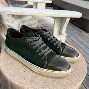 Gröna sneakers från Lanvin - Snygga gröna sneakers från Lanvin i mocka med glansiga detaljer på tån. Skorna har en vit sula och grå snörning som ger en stilren look. Perfekta för en avslappnad men ändå elegant stil. Defekter på tån som man kan se på bild 5. Bara skorna tillkommer. Pris går att diskutera vid snabbt köp. 
