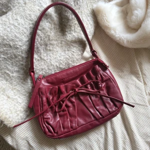 Vinröd axelväska från Coccinelle - Vinröd axelväska i mjukt skinn från Coccinelle. Väskan har snygga rynkade detaljer framtill och en dekorativ knytning. Stängs med dragkedja upptill och har en stadig axelrem. Perfekt storlek för det viktigaste och ger en chic vibe till din outfit.