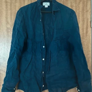 Linneskjorta  - Snygg mörkblå skjorta från H&M Edition i 100% linne. Skjortan har klassisk krage, vita knappar framtill och vid ärmslut. Perfekt för dig som gillar en avslappnad och stilren look. Passar lika bra till jeans som till shorts. Hål i ena ärmen, som ej syns om man kavlar upp. 