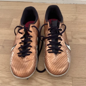 Nike Air Zoom fotbollsskor i koppar med mycket bra skick  - Snygga Nike Air Zoom fotbollsskor i kopparfärg med svarta snören och vita detaljer. Ovandelen har en metallic look och texten 'NIKE ZOOM' syns tydligt. Yttersulan är vit med rosa logga och greppigt mönster, perfekt för inomhusspel. Lätta och smidiga för snabba rörelser.