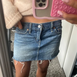 Levi's jeanskjol med fransig kant - Säljer en klassisk blå jeanskjol från Levi's med fransig, rå kant nertill. Kjolen har fem fickor, bälteshällor och knappstängning framtill. Perfekt för dig som gillar en avslappnad och trendig look.