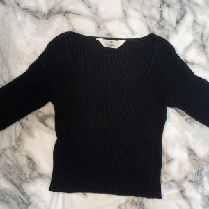 Svart ribbad topp från H&M - En svart ribbad topp från H&M med fyrkantig ringning och lång ärm. Toppen är figurnära och har en enkel, stilren look som passar perfekt till jeans eller kjol. Materialet är stretchigt och bekvämt, vilket gör den lätt att bära hela dagen.