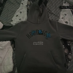 Grå hoodie från Hoodrich med tryck - Säljer en grå hoodie från Hoodrich i storlek M. Tröjan har stor huva, magficka och feta tryck både fram och bak med texten 'From Nothing To Something' och 'Product of the streets'. Snygga blå detaljer på bröstet. Perfekt streetwear-vibe.