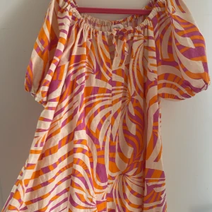 Färgglad kortklänning från Zara - Superfin kortklänning från Zara med färgstarkt mönster i orange, rosa och beige. Klänningen har puffärmar, resår vid halsen och en söt knytning framtill. Ser ut som en klänning men sitter ihop i två korta vida shorts ben men märks inte gör det bara lättare att röra sig i och bekvämare. 