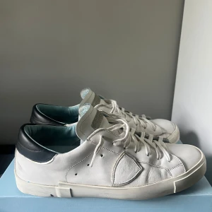 Philippe Model skor - Snygga vita sneakers från Philippe Model Paris med svart häl och ljusblått foder. Skorna har klassisk låg profil, rund tå och snörning. Tillverkade i mjukt skinn med diskret logga på sidan och gummisula för bra grepp. Perfekta för en clean och stilren look.