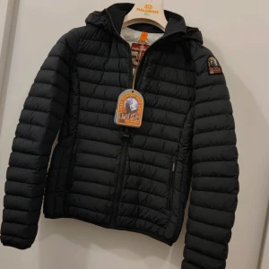 Svart dunjacka från Parajumpers - Snygg svart dunjacka från Parajumpers med huva och dragkedja. Jackan har ett quiltat mönster och en logotyp på ärmen. Perfekt för kyligare dagar.