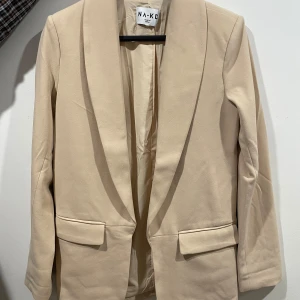 Beige kavaj från NA-KD - Stilren beige kavaj från NA-KD med klassisk krage och två fickor framtill. Rak passform och helfodrad insida. Perfekt för en clean och modern look. Enkel att matcha med både jeans och kostymbyxor.