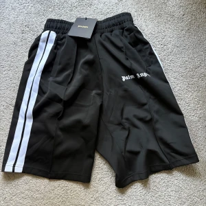 Palm Angels shorts - Svarta shorts från Palm Angels med vita dubbla ränder längs sidorna och logga tryckt på vänster ben. Elastisk midja för skön passform. 