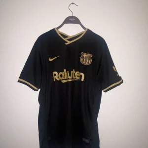 Svart FC Barcelona Nike fotbollströja - Snygg svart FC Barcelona fotbollströja från Nike med guldiga detaljer och tryck  (!ingen spelare!) . Tröjan har korta ärmar, V-ringad krage med guldkant och klubbmärke samt sponsorloggor i guld. Perfekt för dig som älskar Barca eller gillar allmänt.