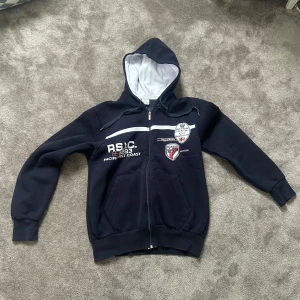 Mörkblå hoodie från Paul & Shark - Säljer en mörkblå hoodie från Paul & Shark med broderade detaljer och tryck både fram och bak. Tröjan har huva med vit insida, dragkedja och två fickor framtill. Perfekt för dig som gillar sportig och avslappnad stil.