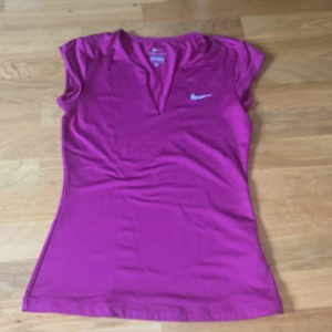 Lila Dri-Fit topp från Nike - Sportig rosa topp från Nike med Dri-Fit-teknologi. Modellen har korta ärmar, v-ringning och en liten vit Nike-logga på bröstet. Perfekt för träning eller tennis. Storlek S.