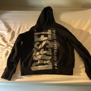 Svart hoodie från Diesel med tryck - Svart hoodie från Diesel med stort vitt och grått tryck över hela framsidan. Klassisk huva och långa ärmar, ribbade muddar och nederkant. Snygg streetstil med tydlig logga och statement-tryck. Perfekt för dig som gillar att sticka ut.
