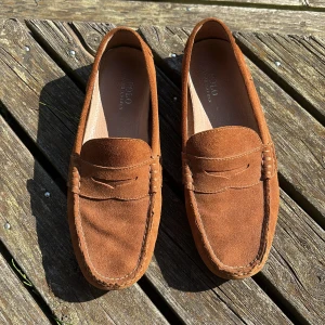 Bruna mockaloafers från Polo Ralph Lauren - Snygga loafers i brun mocka från Polo Ralph Lauren. Klassisk design med rund tå, dekorativ söm och diskret Polo-detalj på sidan. Skorna har platt sula med gummidetaljer undertill för extra grepp. Perfekta för en chill och stilren look.