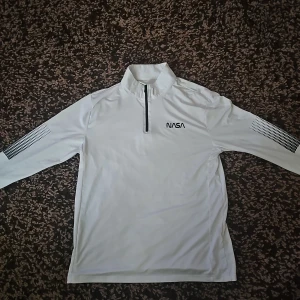 Vit NASA half zip långärmad tröja - Vit långärmad tröja med svart NASA-tryck på bröstet och coola svarta detaljer på ärmarna. Tröjan har en half zip-dragkedja framtill och är gjord i ett mjukt, lätt material som känns sportigt och fräscht. Perfekt för dig som gillar sport och stilren design