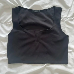 Svart croppad topp från NLY One - Säljer en svart croppad topp från NLY One med breda axelband och en snygg v-formad urringning framtill. Enkel och stilren design som passar perfekt till jeans eller kjol. Toppen är ärmlös och har en tight passform. (Metall) spänne vid v-ringning