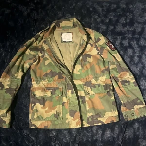 Camouflage parkajacka från Pull&Bear - Snygg parkajacka från Pull&Bear i klassiskt camouflage-mönster med gröna, bruna och beige toner. Jackan har dragkedja, stora fickor framtill och patch på ena ärmen. Perfekt för dig som vill ha en cool streetstyle-look.