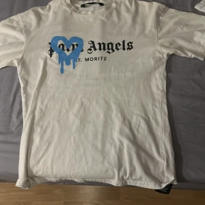 Vit Palm Angels t-shirt med blått hjärta - Behöver bara strykas! Säljer en vit t-shirt från Palm Angels med svart text och ett stort blått droppande hjärta på bröstet. Klassisk passform med korta ärmar och rund hals. Materialet är mjuk bomull, perfekt för en chill och trendig look.