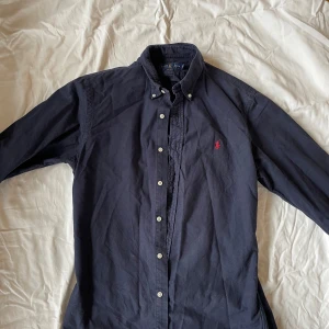 Ralph Lauren Skjorta  - Klassisk skjorta från Ralph Lauren i tidlös mörkblå färg med det ikoniska röda Polo emblemet på bröstet. Modellen är slim fit med buttondown krage. Passar lika bra till vardags som till mer uppklädda tillfällen.  ✔️ 100 % bomull – mjuk och bekväm ✔️ Tidlös design med premiumkänsla ✔️ Original – äkta Ralph Lauren  