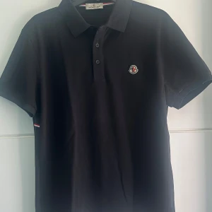 Svart Moncler pikétröja - Svart pikétröja från Moncler med klassisk krage, tre knappar och Moncler-logga på bröstet. T-shirten har korta ärmar och diskreta rödvita detaljer på sidan. Tillverkad i bomull för en skön och stilren känsla.