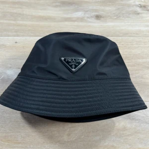 Svart bucket hat från Prada - Stilren svart bucket hat från Prada med ikonisk metallogo framtill. Hatten är tillverkad i glansig syntet och har klassisk form med bred, nedåtlutande brätte. Perfekt accessoar för en trendig look. Rensar garderoben och hittade denna men kan tyvärr inte hitta lådan som kom med