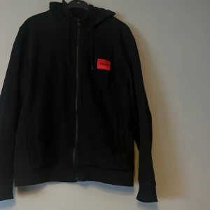 Svart hoodie från Hugo med röd logga - Svart hoodie från Hugo med dragkedja framtill och huva med snörning. På bröstet sitter en röd patch med svart HUGO-tryck. Tröjan har långa ärmar och två fickor framtill. Perfekt för en stilren och avslappnad look.