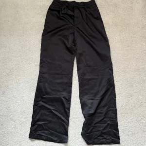 Svarta vida kostymbyxor i satin - Snygga svarta kostymbyxor i satin med hög midja och vida ben. Perfekta för en stilren look. Tillverkade i ett mjukt och bekvämt material som ger en elegant känsla.