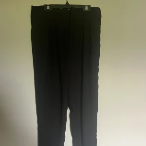 Svarta kostymbyxor från Gina Tricot - Svarta kostymbyxor från Gina Tricot med hög midja och raka ben. Byxorna har klassisk look med pressveck och stängs med dragkedja och hake. Perfekta för en clean och stilren outfit.