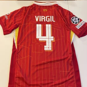 Liverpool Virgil #4 matchtröja Nike - Liverpool FC matchtröja replika med Virgil van Dijk #4 på ryggen. Röd med tunna gula ränder, officiella tryck och Champions League-märke på ärmen. Broderad LFC-logga och Nike swoosh framtill. Tillverkad i lätt och ventilerande polyester. Endast handtvätt gäller