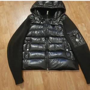 Svart Moncler cardigan - Säljer en svart pufferjacka från Moncler med stickade ärmar och glansig front. Jackan har huva, dragkedja och en snygg ficka med Moncler-logga på ärmen. Perfekt för dig som vill ha en stilren och sportig look med premiumkänsla.