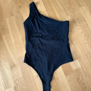 Svart oneshoulder bodysuit - Snygg svart bodysuit med oneshoulder-design och dubbla smala axelband på ena sidan. Tight passform som framhäver siluetten och ger en modern look. Perfekt att styla med jeans eller kjol för en trendig outfit.
