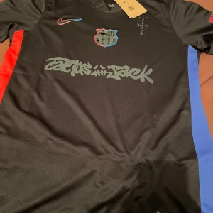 FC Barcelona x Cactus Jack Nike tröja - Snygg svart FC Barcelona fotbollströja i samarbete med Cactus Jack och Nike. Tröjan har korta ärmar, tryck i rött och blått på sidorna, samt unika detaljer som Cactus Jack-grafik och UNHCR-logga. Materialet är lätt och andas, perfekt för match eller träning.
