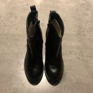 Svarta boots i skinn med klack - Stilrena svarta boots i äkta skinn med rund tå och markerad klack. Dragkedja på insidan för enkel påtagning. Perfekta för dig som vill ha en klassisk och tidlös look med lite edge. Tillverkade i Portugal.
