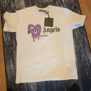 Vit Palm Angels t-shirt med lila hjärta - Snygg vit t-shirt från Palm Angels med svart tryck och ett stort lila hjärta med drip-effekt på bröstet. Klassisk passform och korta ärmar, perfekt för dig som vill sticka ut med streetstyle. Mjuk bomullskvalitet och cool Aspen-print.