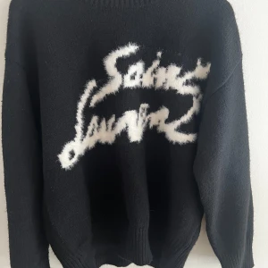 Svart Saint Laurent stickad tröja - Svart stickad tröja från Saint Laurent med vit, fluffig logga framtill i cool graffiti-stil. Tröjan har rund hals, långa ärmar och ribbade muddar. Perfekt för dig som vill sticka ut med en statement piece och gillar exklusiva märken.