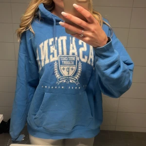 Blå hoodie från Pull&Bear - Säljer en blå hoodie från Pull&Bear med vit tryckt text och emblem på bröstet. Tröjan har huva med snörning, stor magficka och långa ärmar. Perfekt för dig som gillar en avslappnad och sportig stil. Har en fläck vid loggan som inte går bort. 