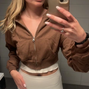 Brun croppad zip-hoodie från H&M  - Säljer en brun croppad jacka från H&M Divided med dragkedja framtill och stora fickor. Jackan har långa ärmar och är gjord i mjuk bomull, perfekt för en chill och avslappnad look. Passformen är loose och den funkar grymt till jeans eller kjol. Så mysig.