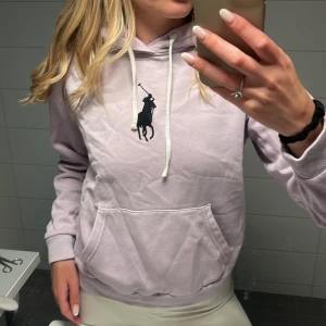 Ljuslila hoodie från Polo Ralph Lauren - Superfin ljuslila hoodie från Polo Ralph Lauren med stor svart broderad logga på bröstet. Tröjan har huva med vita snören, känguruficka och ribbade armmuffar. Skulle säga att färgen är lite mer lila i verkligheten. Storlek M men skulle mer säga att det är en Xs/S om man vill ha den mer oversized. 