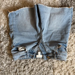 Ljusblå jeansshorts från H&M - Säljer ett par ljusblå jeansshorts från H&M i klassisk denim med råa, fransiga benslut. Shortsen har femficksdesign, bälteshällor och dragkedja med knapp. Perfekta för en chill sommarlook.