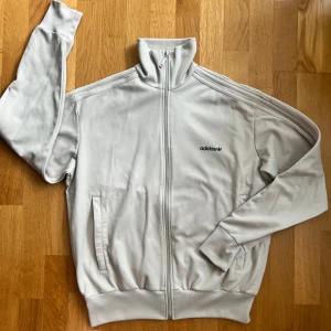 Beige Adidas track jacket XL - Snygg vit Adidas track jacket med klassiska tre ränder längs ärmarna och broderad logga på bröstet. Jackan har hög krage, hel dragkedja och två fickor framtill. Storlek XL men passar som Large