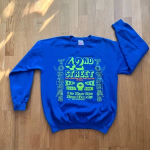 Milk Crate - Blå 42nd Street sweatshirt i medium - Cool blå sweatshirt med stor gul och orange 42nd Street-tryck framtill. Tröjan har rund hals, långa ärmar och är gjord i en mjuk bomulls- och polyestermix. Perfekt statement piece för dig som gillar retro vibes och färgstarka prints.