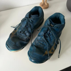 Blå sneakers från Mizuno med snörning - Säljer ett par blå sneakers från Mizuno med guldiga detaljer och klassisk snörning. Ovandelen är i mesh och syntet för bra andningsförmåga. Skorna har rund tå och platt sula, perfekta för träning eller vardagsstil. De är rätt slitna. De har använts som handbollsskor så där är några fläckar på de från klister. 
