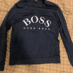 Mörkblå Hugo Boss sweatshirt S - Snygg mörkblå sweatshirt från Hugo Boss med stort vitt BOSS-tryck på bröstet. Tröjan har rund hals, långa ärmar och ribbade muddar. Perfekt för dig som gillar stilrena och klassiska plagg med tydlig logga.