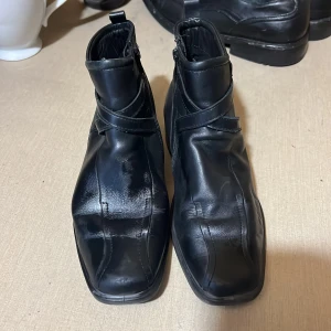 Svarta skinnstövletter från Ecco - Snygga svarta stövletter från Ecco i äkta skinn med fyrkantig tå och låg klack. Skorna har dragkedja på insidan och ett dekorativt spänne runt ankeln. Perfekta för dig som gillar stilrena och klassiska boots med modern touch.