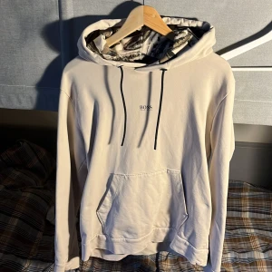 Beige hoodie från Hugo Boss med mönstrad huva - Snygg beige hoodie från Hugo Boss med svart logga på bröstet. Huvan har ett unikt camouflageinspirerat mönster i beige, svart och brunt. Tröjan har känguruficka, svarta snören och är tillverkad i mjuk bomull. Perfekt för dig som vill ha en stilren men ändå unik look.